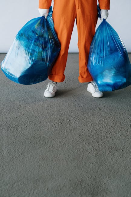 A person dressed in orange work overalls and white sneakers is standing on a grey carpeted floor, holding two large blue plastic rubbish bags filled with waste materials. The individual’s hands are